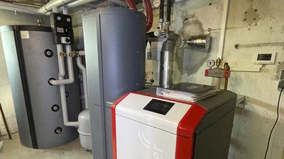 Im Hintergrund sieht man einen großen, grauen Warmwasserspeicher neben einem kompakten Heizgerät mit rotem Akzent.
