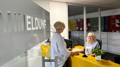Eine blonde Frau mit Brille lächelt am Empfangstresen, während eine andere Frau mit kurzem Haar ihr gegenübersteht.