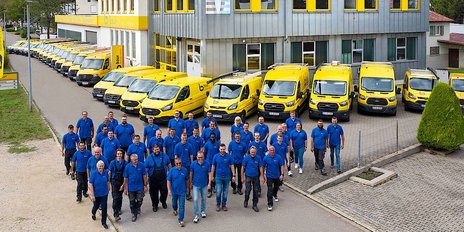 Eine Gruppe von Männern in blauen T-Shirts marschiert vor einem modernen Gebäude mit gelben Lieferwagen.