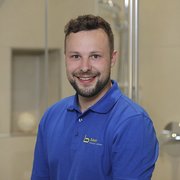 Professionelles Porträt eines freundlichen Handwerkers im Badezimmer Ein lächelnder Mann in einem blauen Poloshirt sitzt vor einer modernen Badezimmerumgebung mit hellen Fliesen.