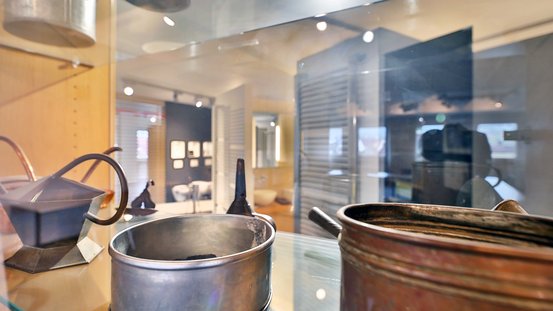 In einem Ausstellungsraum sind verschiedene antike Metallbehälter zu sehen, die auf einem Glasregal präsentiert werden.