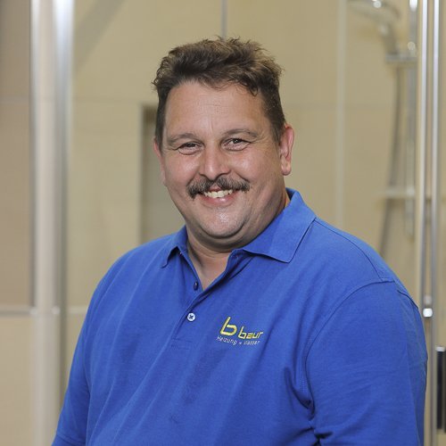 Ein lächelnder Mann mit einem Schnurrbart trägt ein blaues Poloshirt und steht vor einer modernen Badezimmerumgebung.