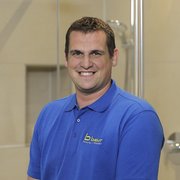 Professioneller Handwerker in blauer Arbeitskleidung lächelt freundlich Ein lächelnder Mann in einem blauen Poloshirt steht vor einer modernen Duschkabine und strahlt Freundlichkeit aus.