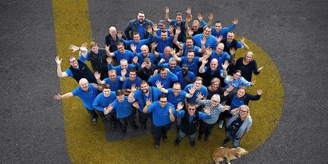 Eine große Gruppe von Menschen in blauen T-Shirts steht auf einem grauen Boden und hebt fröhlich die Hände in die Luft.