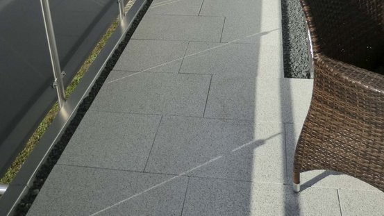 Ein gepflasterter Balkon mit grauen Fliesen und einem geflochtenen Stuhl, der im Sonnenlicht steht.