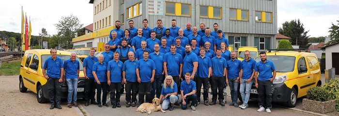 Eine große Gruppe von Mitarbeitern in blauen T-Shirts steht vor einem modernen Gebäude mit gelben Fenstern und gelben Fahrzeugen.