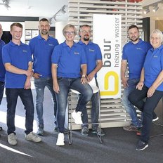 Eine Gruppe von zwölf Personen in blauen T-Shirts steht und sitzt in einem modernen Büro, lächelnd und freundlich.
