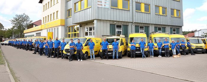Eine Gruppe von Mitarbeitern in blauen Hemden steht vor einem Gebäude mit gelben Akzenten und mehreren gelben Fahrzeugen.