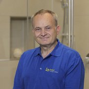 Professioneller Handwerker in blauer Arbeitskleidung lächelt freundlich Der Mann mit kurzem Haar und einem freundlichen Lächeln trägt ein Poloshirt mit dem Logo von Baur und posiert in einem Badezimmer.